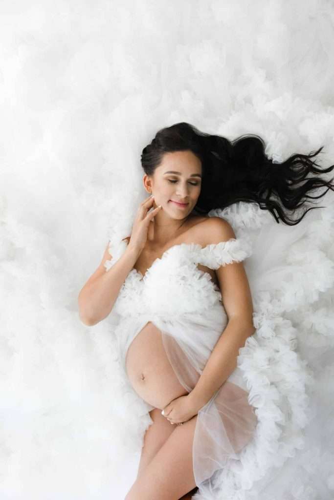 Pregnant woman in a white ruffled dress lies on her back with eyes closed, touching her face and displaying her baby bump against a soft white background.