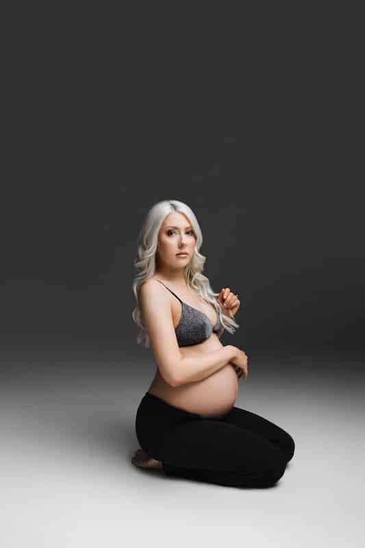 A pregnant woman with long blonde hair kneels on the floor, wearing a gray bra and black pants, against a dark gradient background.