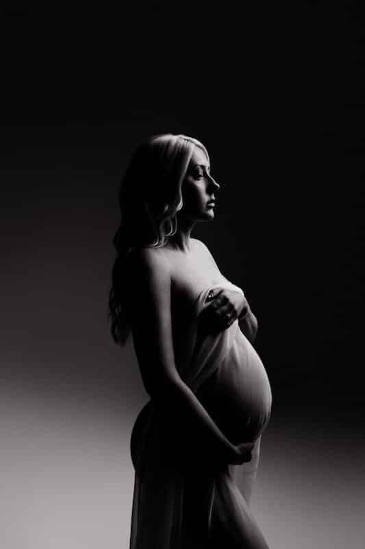 A pregnant woman stands in profile, draped in sheer fabric, with dramatic lighting creating a shadowed background.
