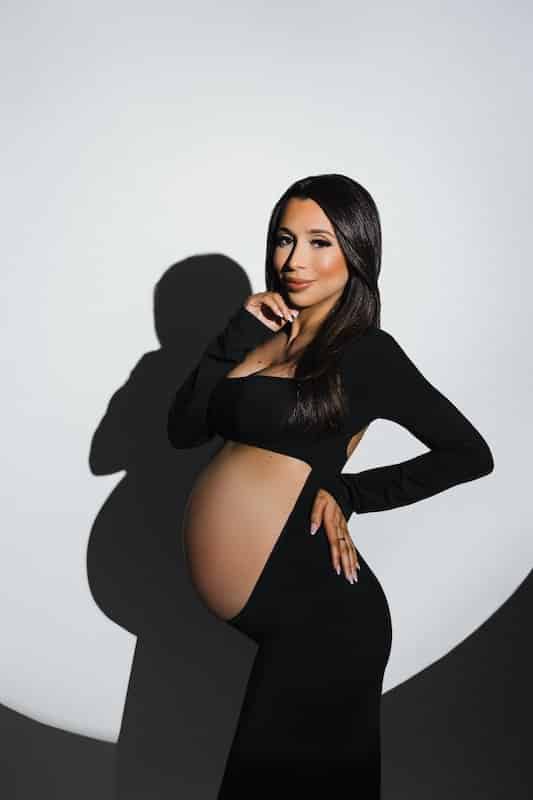 A pregnant woman in a long black dress poses against a white background with a circular spotlight effect, exposing her baby bump.