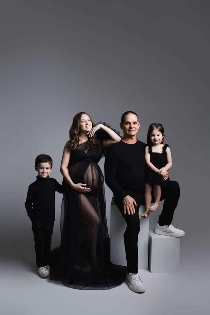 A family of four poses in a studio; the mother is visibly pregnant and wearing a sheer black dress, while the father and two children, a boy and a girl, wear black outfits and white shoes.