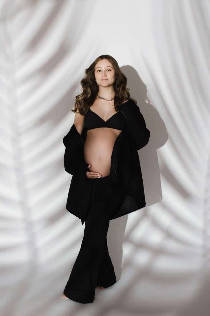 A pregnant woman in black pants and a jacket with an exposed belly stands barefoot against a white background with leaf shadows.