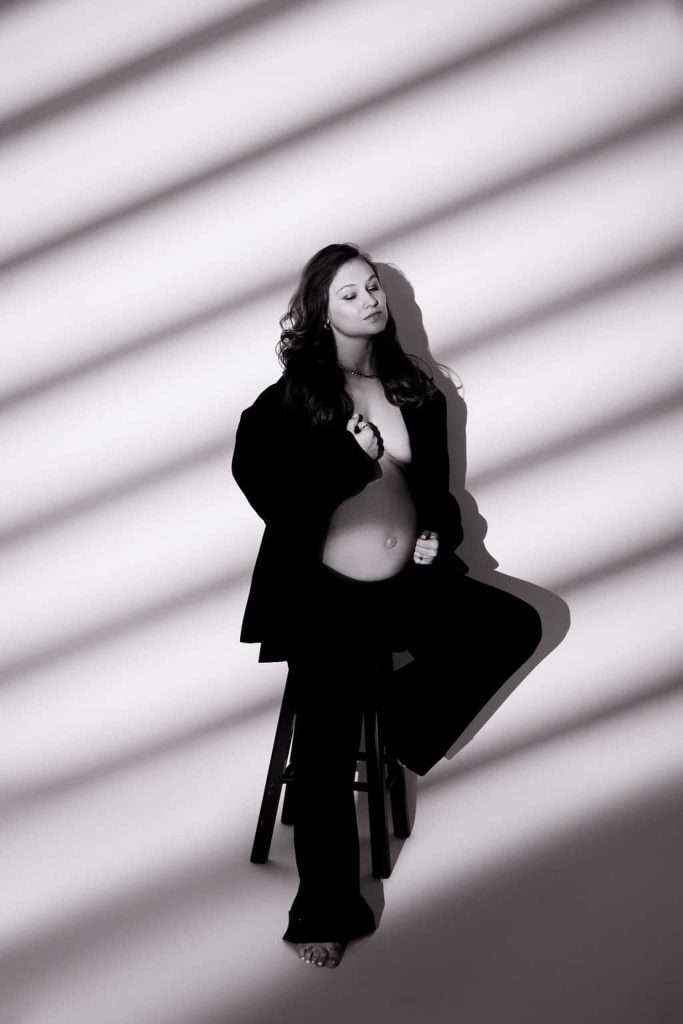 A pregnant woman sits barefoot on a stool, partially dressed in an open jacket, with striped shadows cast across her and the wall.