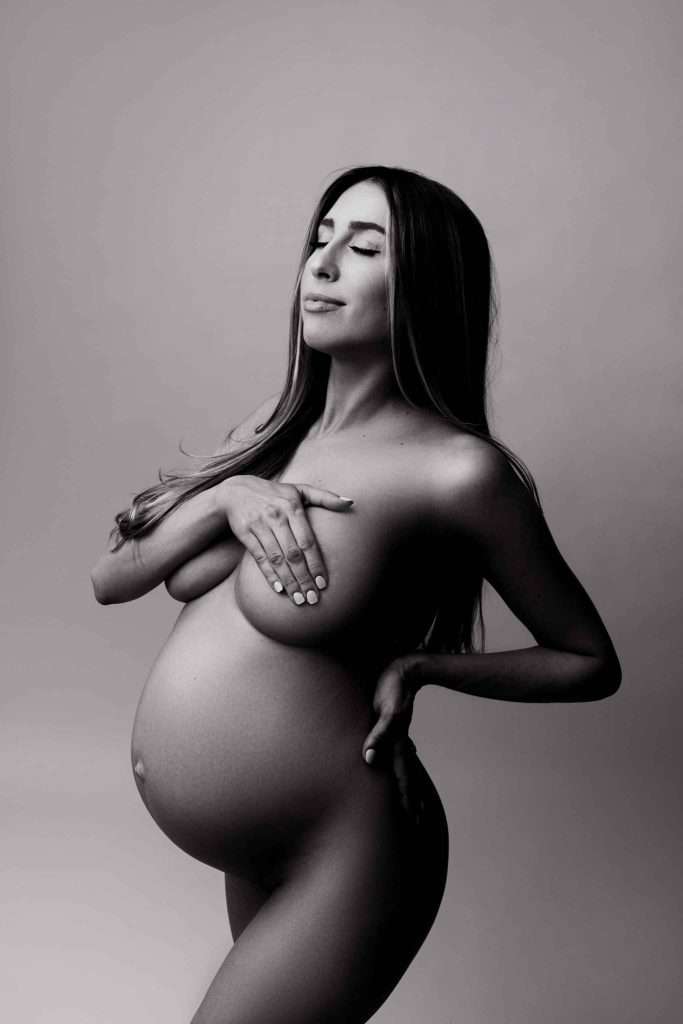 A pregnant woman poses nude in a studio, covering her chest with one hand and resting the other on her hip. The image is in black and white.