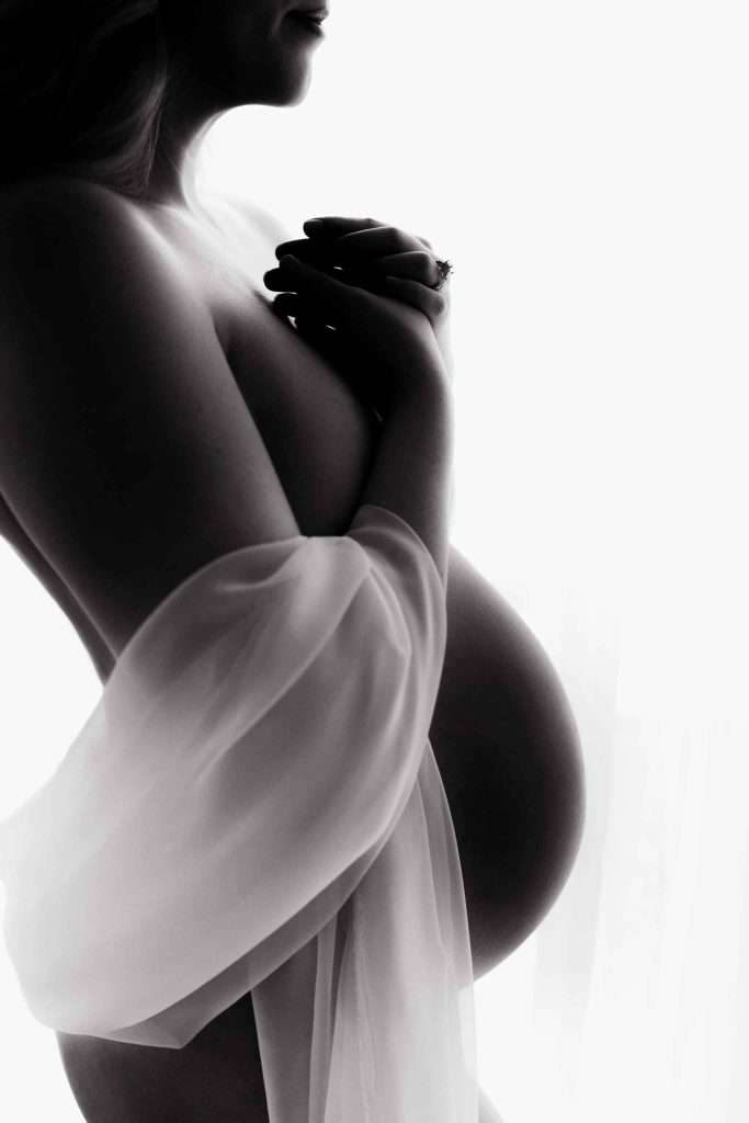 Black and white photo of a pregnant woman in profile, draped in sheer fabric, holding her hands near her chest.