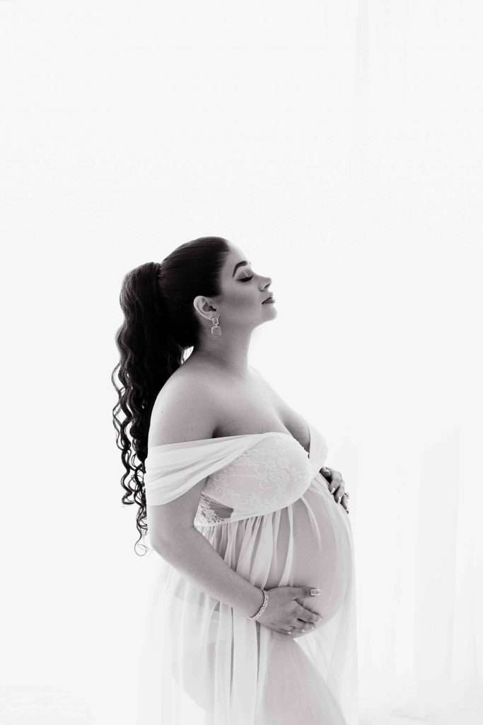 A pregnant woman in a sheer, off-shoulder dress stands in profile, holding her belly and looking upward with a calm expression against a white background.