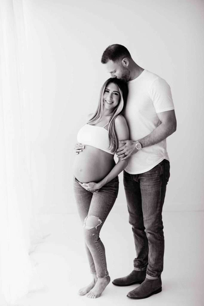 Pregnant woman in a white top and ripped jeans stands barefoot, smiling, while a man in jeans and a white shirt stands behind her, kissing her forehead and holding her belly.