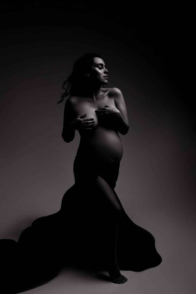 A pregnant woman poses in dramatic lighting, partially draped in dark fabric, covering her chest with her hands.