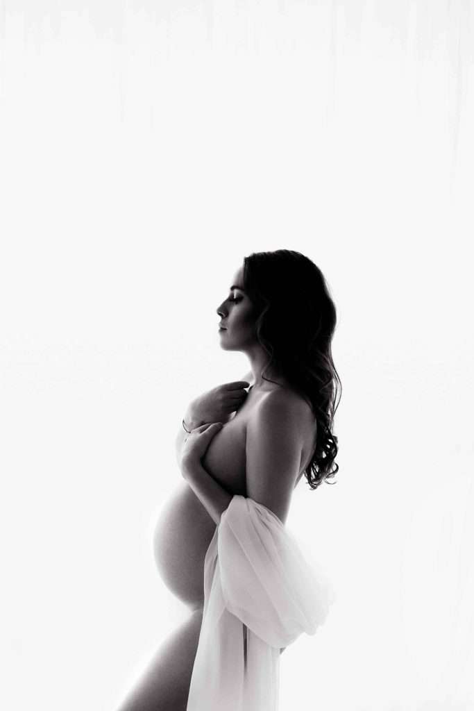 Black and white side profile of a pregnant woman with long hair, draped in a sheer fabric, holding her hand to her chest against a white background.