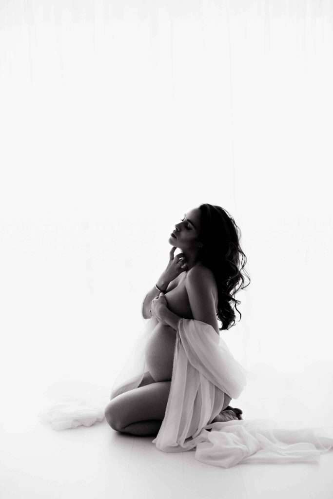 A pregnant woman draped in sheer fabric kneels on the floor, looking upward with one hand near her neck and the other on her belly, against a bright white background.