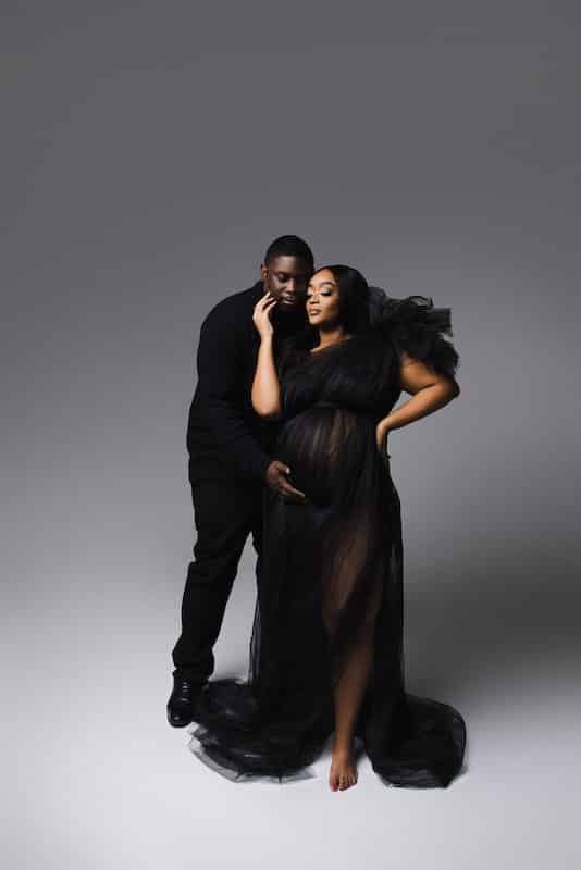 A man and a pregnant woman pose together in a studio. The woman wears a sheer black dress, and the man stands behind her with his hand on her baby bump.