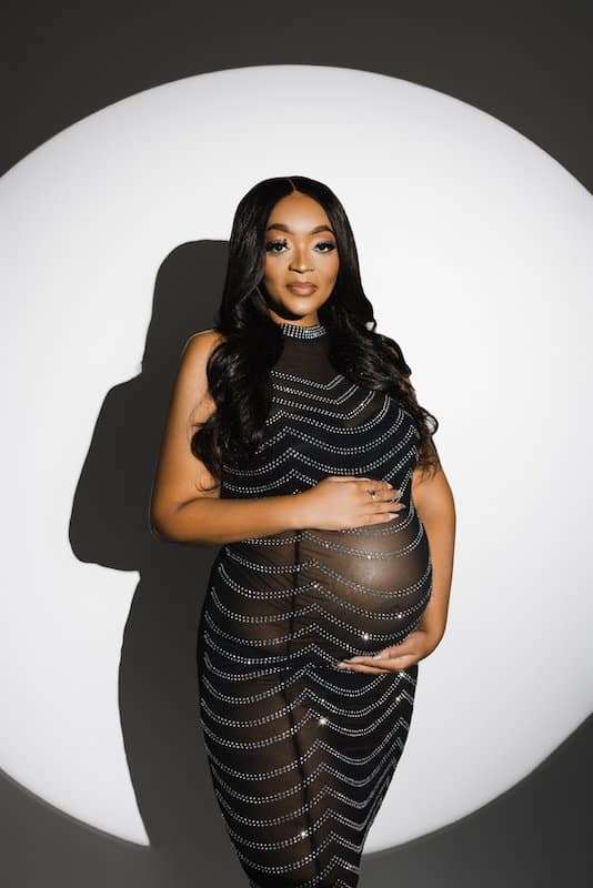 A pregnant woman in a sheer, patterned dress stands in front of a circular white backdrop, cradling her belly with both hands.