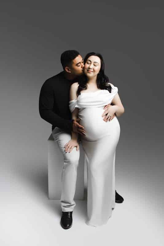 A man kisses a pregnant woman on the cheek as they sit together; she wears a white dress and holds her belly, while he rests his hands on her abdomen.