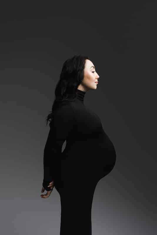 A pregnant woman in a black turtleneck dress stands in profile against a dark gray background, looking forward with her hands behind her back.