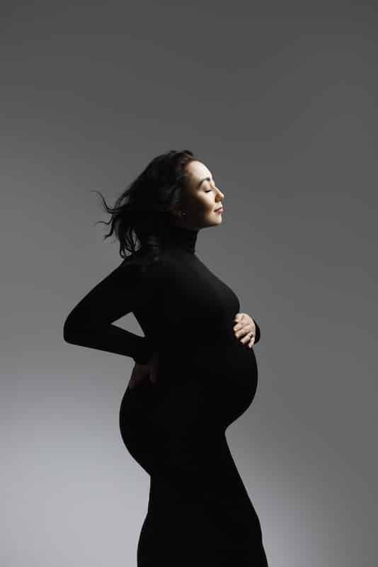 Pregnant woman in a black turtleneck dress stands in profile against a gray background, one hand on her lower back and the other on her belly.