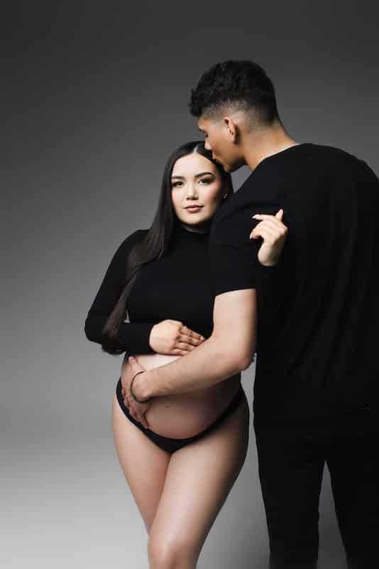 A pregnant woman in black poses with a man who kisses her forehead. She holds her belly and both wear black clothing against a gray background.