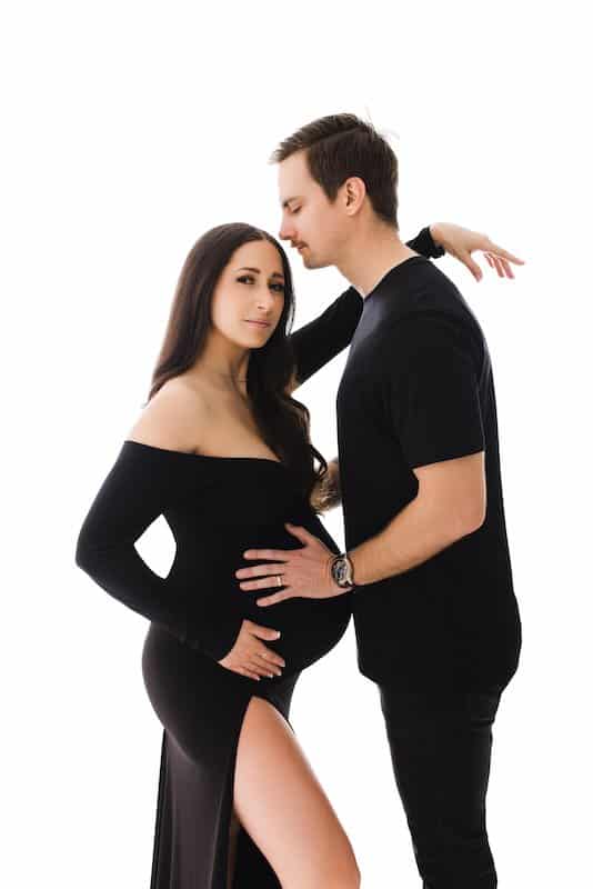Pregnant woman in a black dress stands beside a man in black clothing. He touches her belly and leans in with eyes closed. White background.