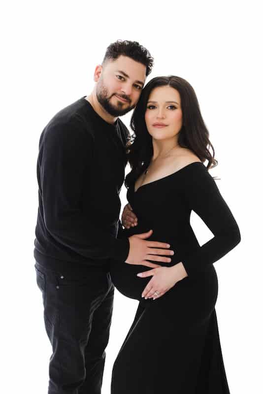 A man and a woman in black outfits stand together against a white background. The man gently places his hand on the woman’s pregnant belly.
