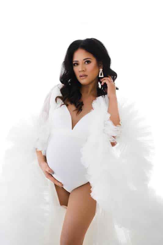 Pregnant woman in a white bodysuit and sheer robe stands against a white background, holding her belly and looking directly at the camera.