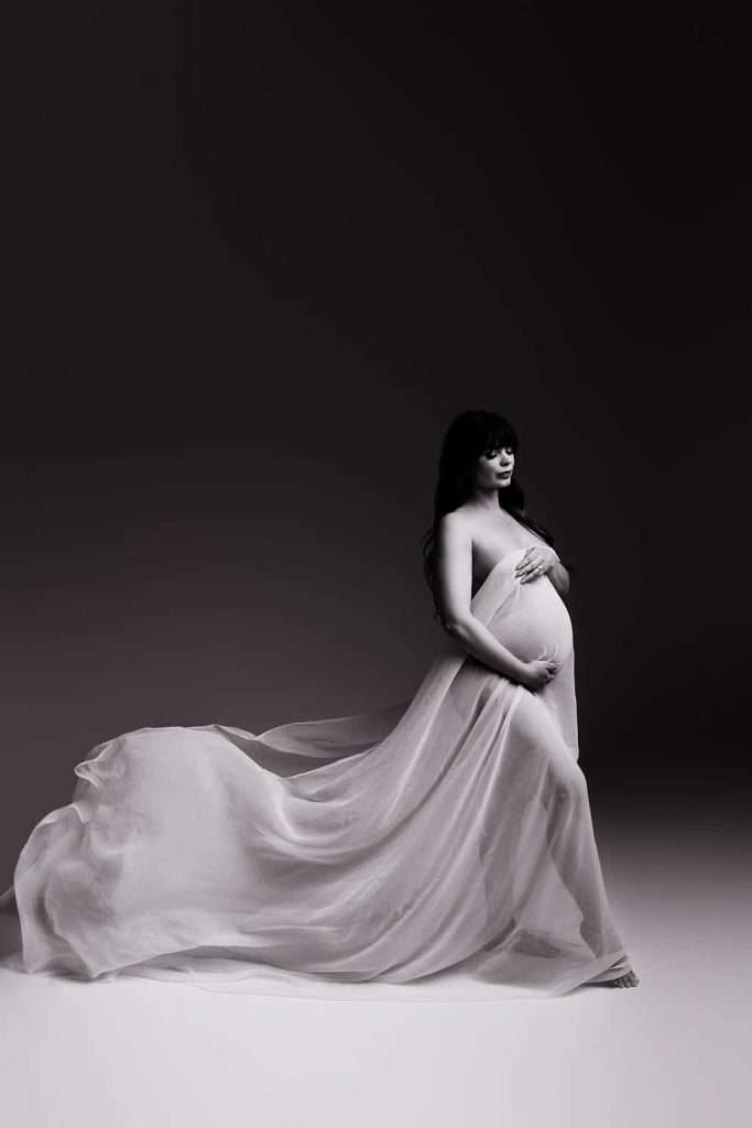 Pregnant woman standing barefoot, draped in flowing sheer fabric, holds her belly and looks forward. The photo is in black and white with a dark gradient background.