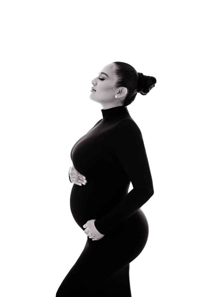 A pregnant woman in a black fitted dress stands in profile against a white background, one hand on her belly and the other on her chest.