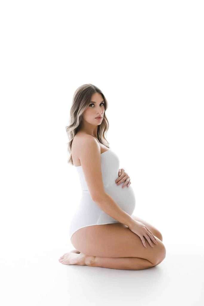 Pregnant woman in a white bodysuit kneeling on the floor, touching her belly, and looking at the camera against a white background.