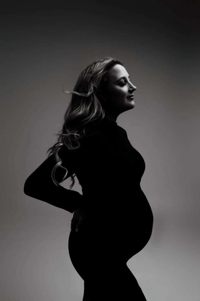 Black and white side profile of a pregnant woman standing with one hand on her lower back, wearing a long-sleeved fitted dress.
