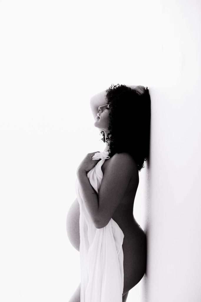 A pregnant woman stands in profile against a wall, draped with a white cloth, with one hand on her head and eyes closed.