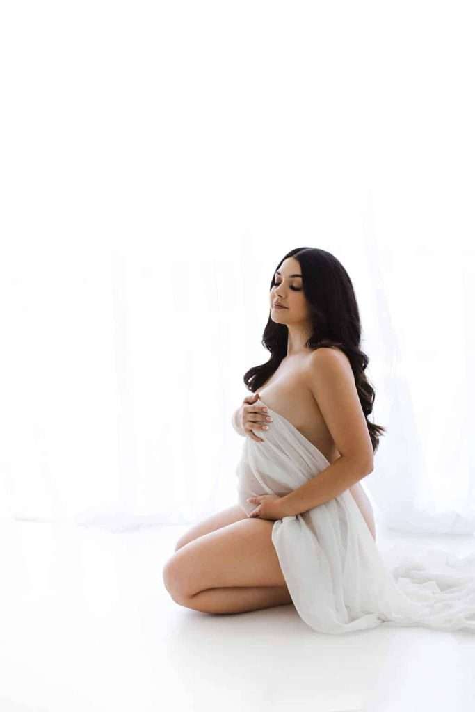 A pregnant woman kneels on the floor, draped in a white fabric, with one hand on her belly and the other covering her chest, against a bright white background.