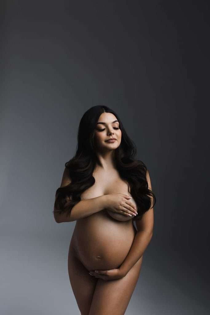 A pregnant woman stands nude, covering her chest with one hand and her belly with the other, against a neutral background.