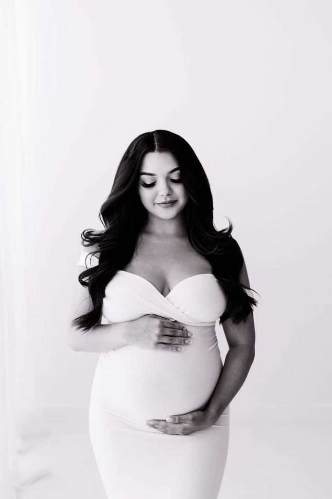 Black and white photo of a pregnant woman in a fitted dress, standing and gently holding her belly with both hands, looking down with a calm expression.