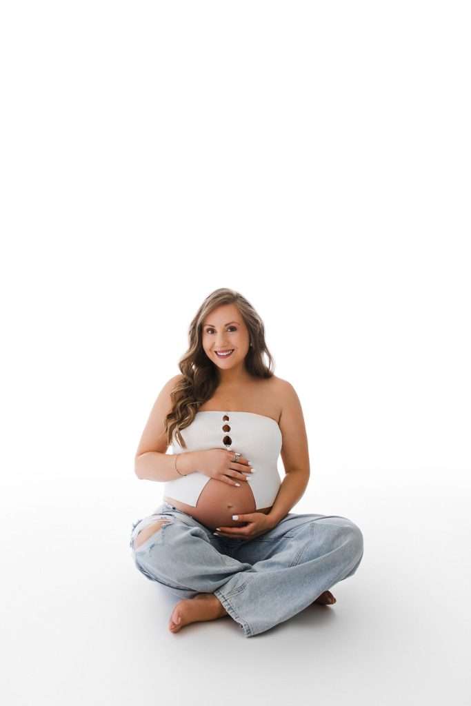 Pregnant woman in a white top and ripped jeans sits cross-legged on a white floor, smiling and gently holding her belly with both hands.