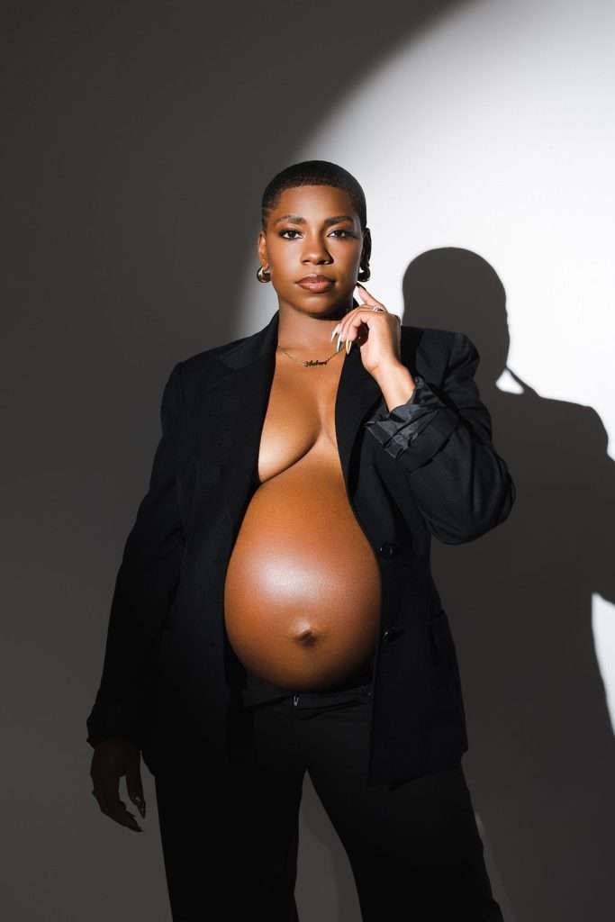 A pregnant person poses confidently in a black blazer and pants, exposing their bare belly, against a white background with dramatic lighting and shadow.