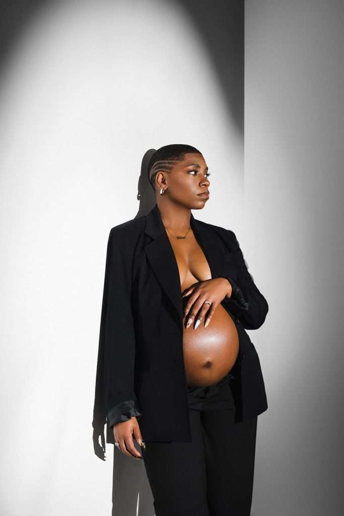 A pregnant woman in a black suit poses in a well-lit corner, one hand on her bare belly, looking to the side with a calm expression.
