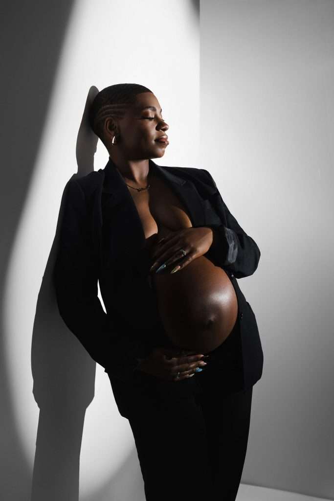 A pregnant person in a black suit jacket stands against a white wall, cradling their bare belly and looking to the side in dramatic lighting.