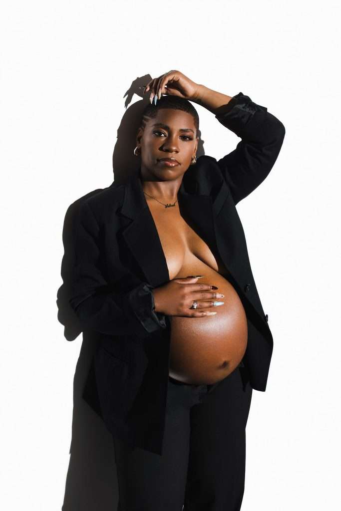 A pregnant woman in a black blazer and pants poses confidently against a white background, with one hand on her head and the other resting on her exposed belly.