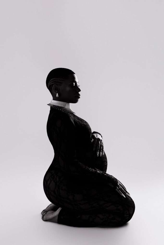 A pregnant woman in a fitted black lace outfit kneels on the floor, her hand on her belly. She wears a necklace and earrings, with a short, styled haircut against a plain background.