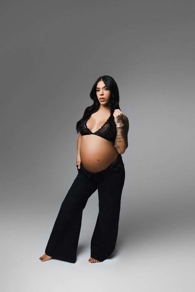 A pregnant woman stands barefoot in black pants and a black lace bralette against a plain gray background, looking directly at the camera.