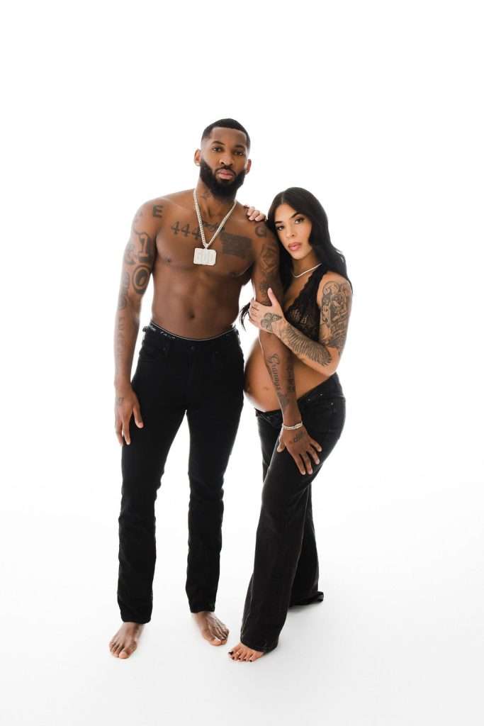 Shirtless man and pregnant woman wearing black pants stand together against a white background, showing their tattoos.