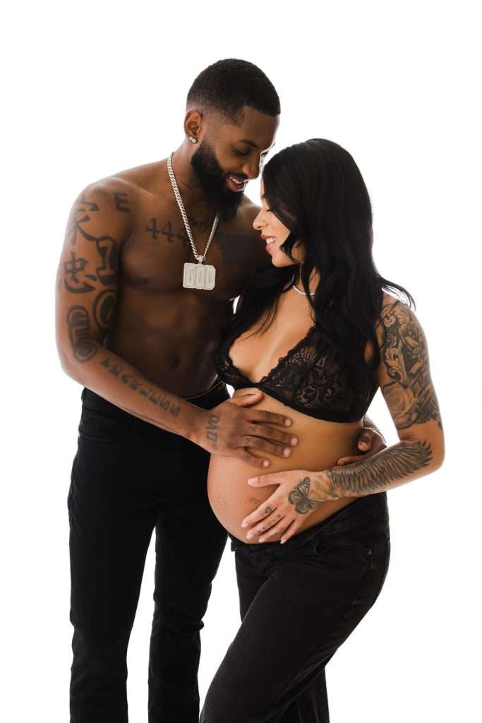 A shirtless man and a woman in a black lace bra stand together. Both have tattoos and are touching the woman’s pregnant belly, looking at each other, on a white background.