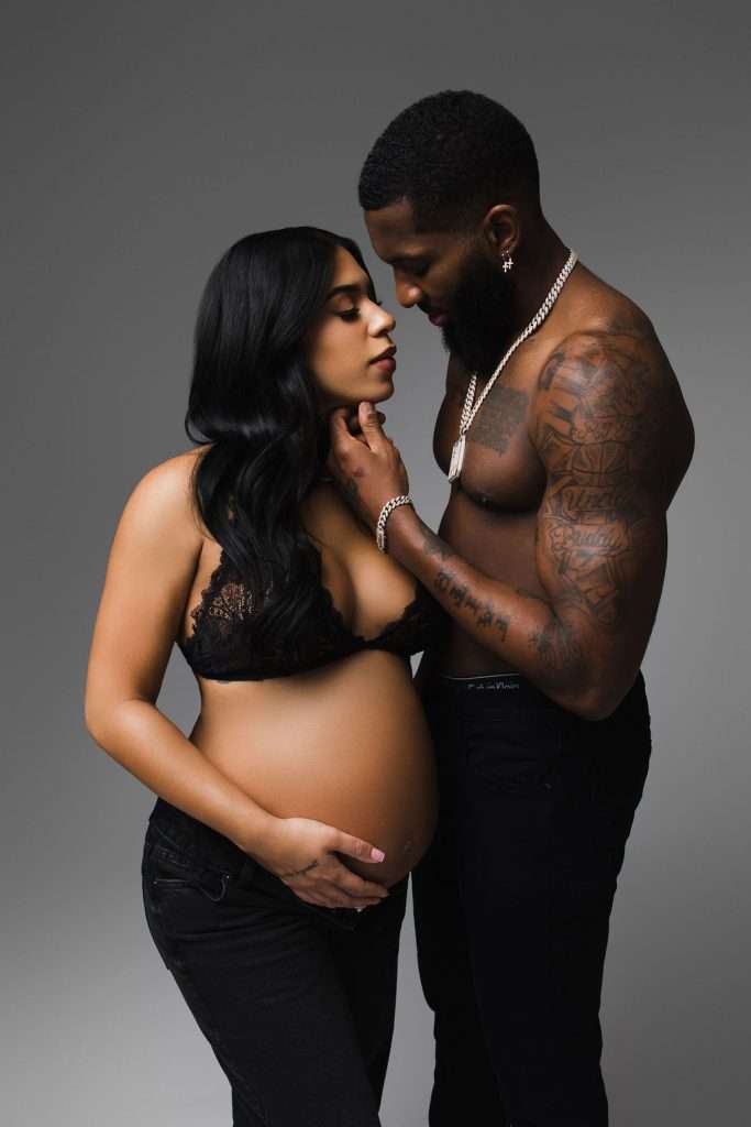 A pregnant woman in a black lace top stands facing a shirtless man with tattoos. They are embracing, with her hand on her belly and his hand gently touching her face.