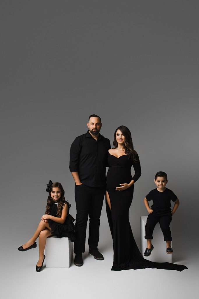 A family of four poses in a studio. The father and pregnant mother stand together, while their young daughter and son sit on white blocks, all dressed in black outfits against a gray background.