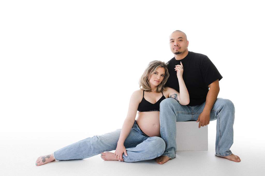 Pregnant woman in a black bra and jeans sits on the floor beside a man in a black shirt and jeans, against a white background.