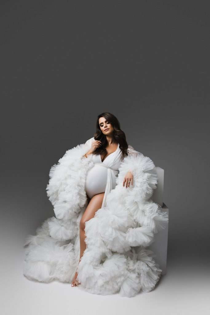 Pregnant woman wearing a voluminous white ruffled robe sits on a white block against a gray background.