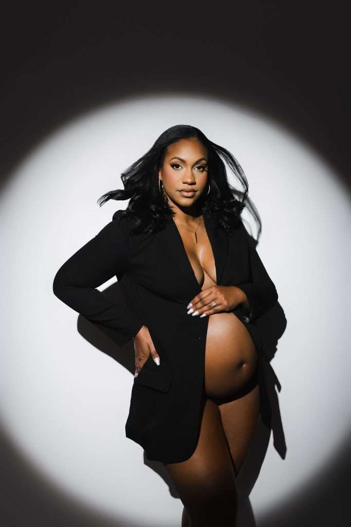 A pregnant woman wearing a black blazer poses confidently under a spotlight, with her hand on her hip and her baby bump exposed.