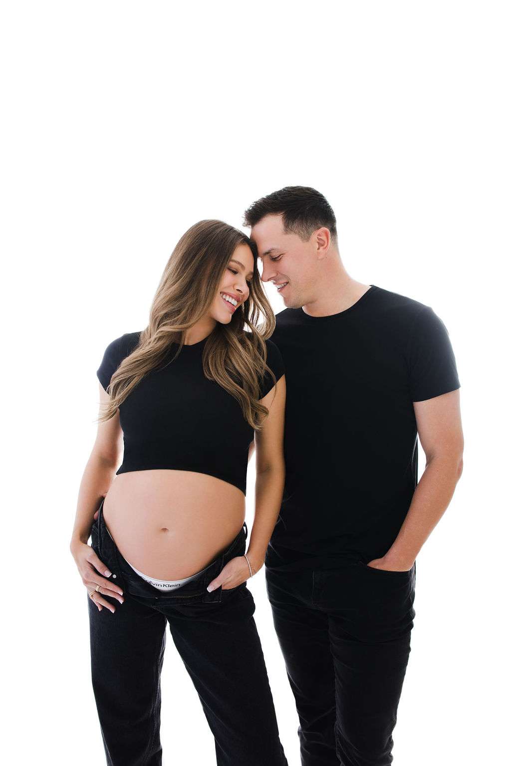 Pregnant woman and man stand together wearing black outfits. The woman shows her bare belly and both are smiling, looking at each other, against a white background.