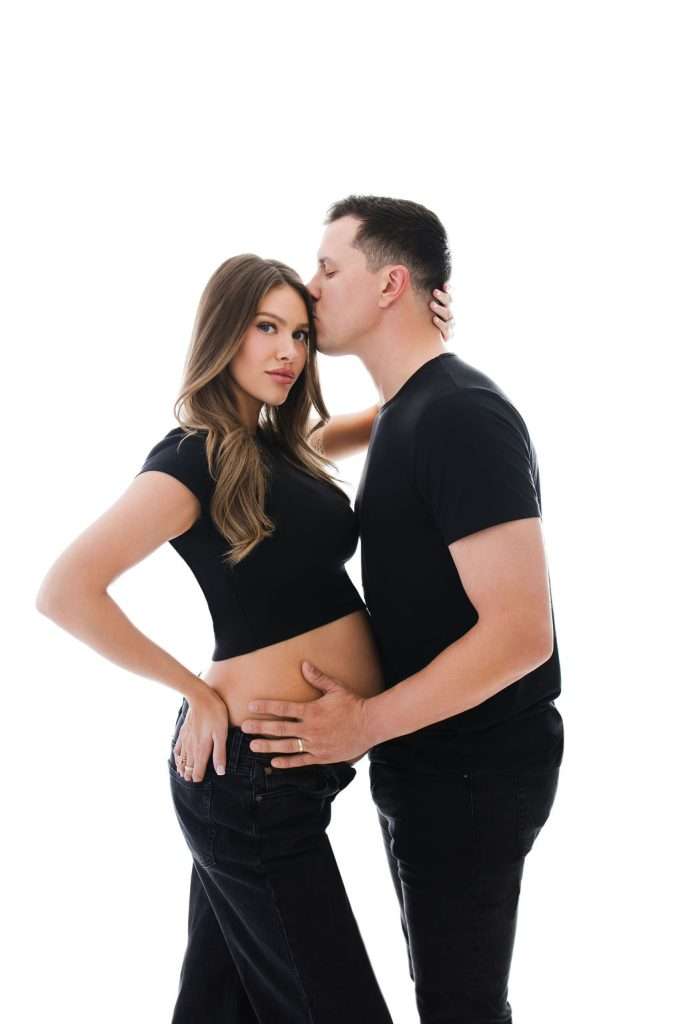 A man kisses a pregnant woman’s forehead while gently holding her belly. Both wear black shirts and jeans, standing against a plain white background.