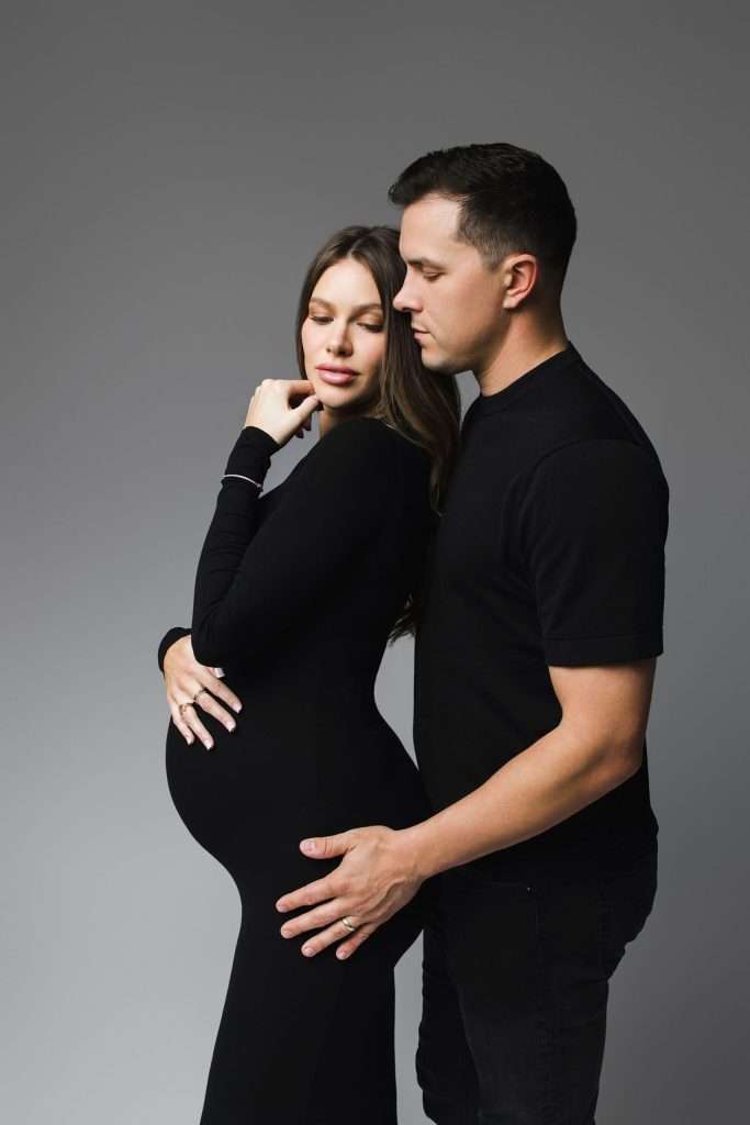 A pregnant woman in a black dress stands next to a man in a black shirt, both with neutral expressions against a plain gray background.