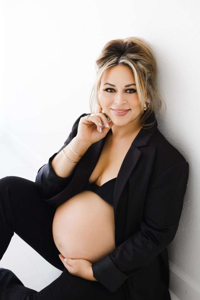 A pregnant woman in a black blazer and bra sits on the floor, smiling while holding her belly with one hand and touching her chin with the other.