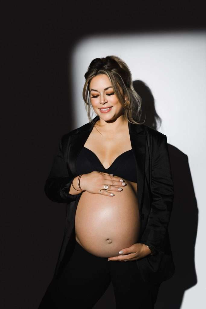 A pregnant woman wearing a black bra and blazer stands against a white and dark background, smiling and holding her exposed belly with both hands.